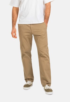 Man wearing beige casual pants, white sneakers, and a white short-sleeve shirt, standing with one hand in pocket against a plain background.