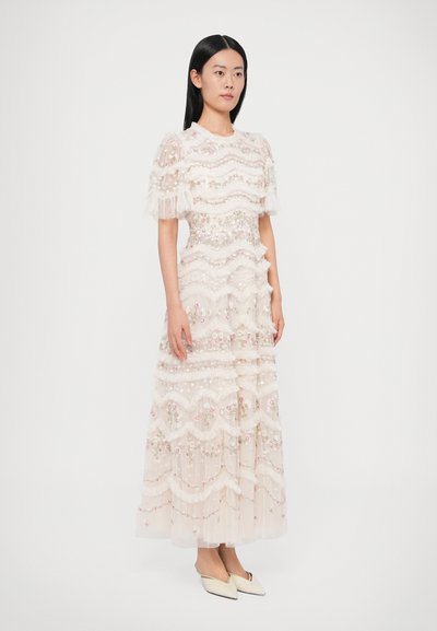 Dlhé krémové šaty z vrstvenej, textúrovanej tkaniny s kvetinovou výšivkou a volánikovými rukávmi. K outfitu ladia špicaté béžové topánky.