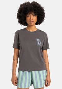 Gray cotton t-shirt with a crew neck and short sleeves, featuring a colored graphic on the left chest pocket area. Paired with striped shorts.