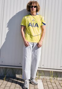 Gul Nike fodboldtrøje med blå detaljer, prydet med "AIA" logo. Bæret med lyse denim jeans og grå sneakers. Udendørs miljø med skygger.