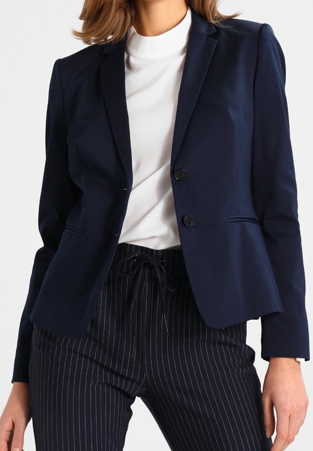 Granatowa marynarka o dopasowanym kroju, z pojedynczym zapięciem na guzik i dwoma przednimi kieszeniami. Nosi się ją na białym golfie, w zestawieniu z spodniami w prążki.