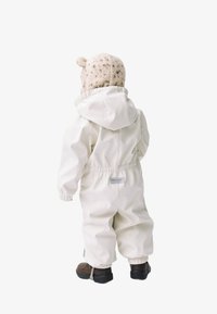 Combinaison blanche imperméable tout-en-un avec une capuche, des poignets élastiques et une taille froncée. Associée à un bonnet en tricot beige avec des oreilles et des bottes marron.
