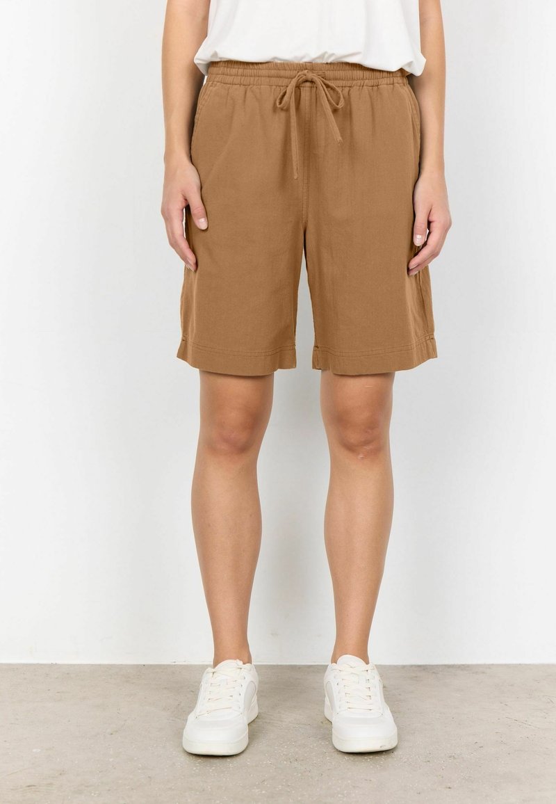 Personne portant un short marron à cordon de serrage jusqu'aux genoux, des baskets blanches et un haut blanc, debout devant un fond blanc uni.