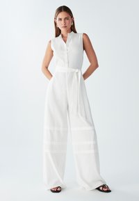 Mono de lino blanco con cuello de camisa, botonera frontal y piernas anchas con acentos de rayas horizontales. Cinturón de tela atado en la cintura.