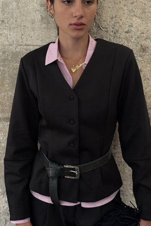 Jeune femme portant un blazer noir boutonné avec ceinture, col de chemise rose visible, collier cœur en or, debout contre un mur en pierre texturé.
