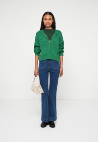 Zöld kötött, cipzáras pulóver texturált mintával, amelyet kék, bő szárú farmerral és világos bézs táskával párosítottak. Fekete lábbeli egészíti ki az öltözéket.