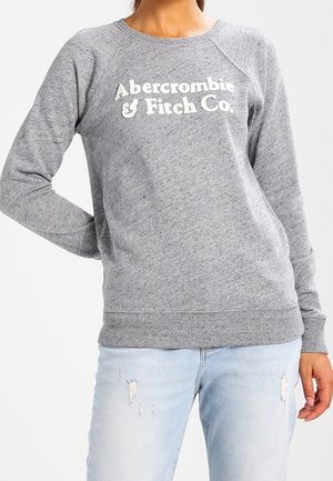 Personne portant un sweat gris Abercrombie & Fitch Co. et un jean bleu clair déchiré, debout avec une main sur la hanche.