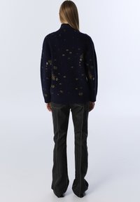 Navy sweater with a high collar, featuring gold star patterns, paired with black flared jeans accented by white stitching, worn from the back.