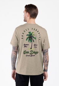 Hombre con tatuajes que lleva una camiseta beige con una palmera y el texto "Verano Paraíso, San Diego Ocean Beach, Desde 1994" en la parte de atrás.