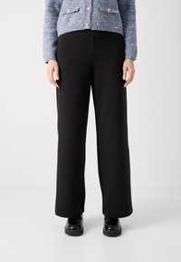 Široké černé kalhoty vyrobené z měkké tkaniny, s hladkým povrchem a volným střihem, kombinované s černými masivními botami.