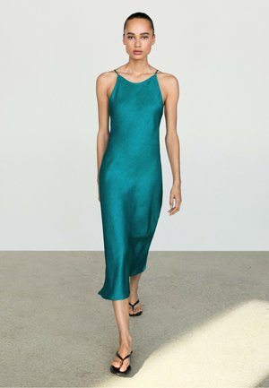 Donna che cammina su un pavimento neutro, indossando un vestito midi senza maniche color teal con sottili spalline e sandali neri.