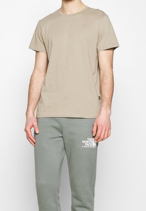 Mężczyzna w beżowej koszulce z krótkim rękawem i szarych spodniach dresowych z logo "The North Face" i współrzędnymi na lewym udzie.