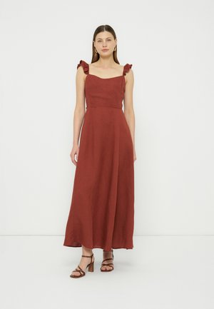 Woman wearing a sleeveless rust-red maxi dress with ruffle straps and brown heeled sandals, standing against a plain white background.