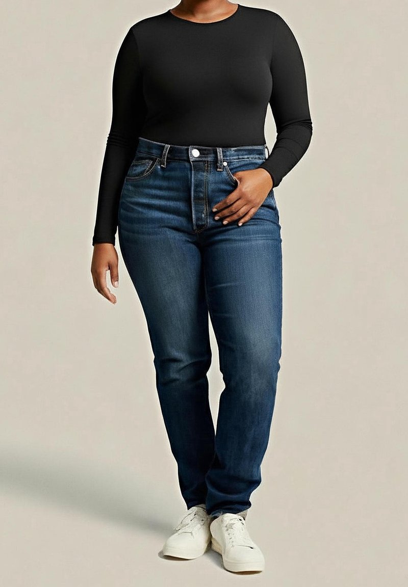 Kvinde iført en sort langærmet top, mørkeblå jeans med høj talje og hvide sneakers, stående med den ene hånd på hoften.