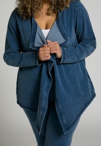 Giacca in denim di colore blu scuro con colletto a scialle e orlo asimmetrico. Maniche lunghe con dettagli a bottoni. Tessuto texturizzato con un design casual.