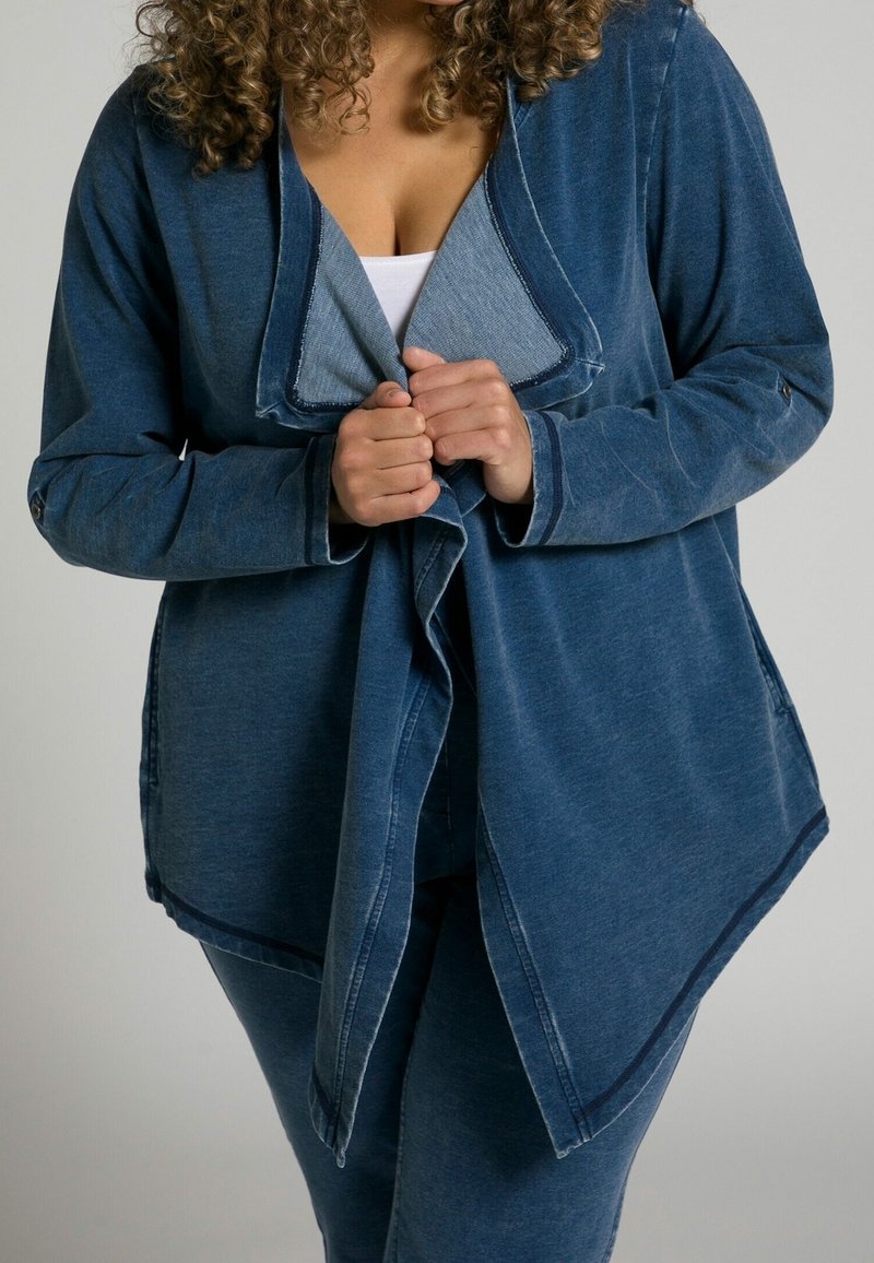 Giacca in denim di colore blu scuro con colletto a scialle e orlo asimmetrico. Maniche lunghe con dettagli a bottoni. Tessuto texturizzato con un design casual.