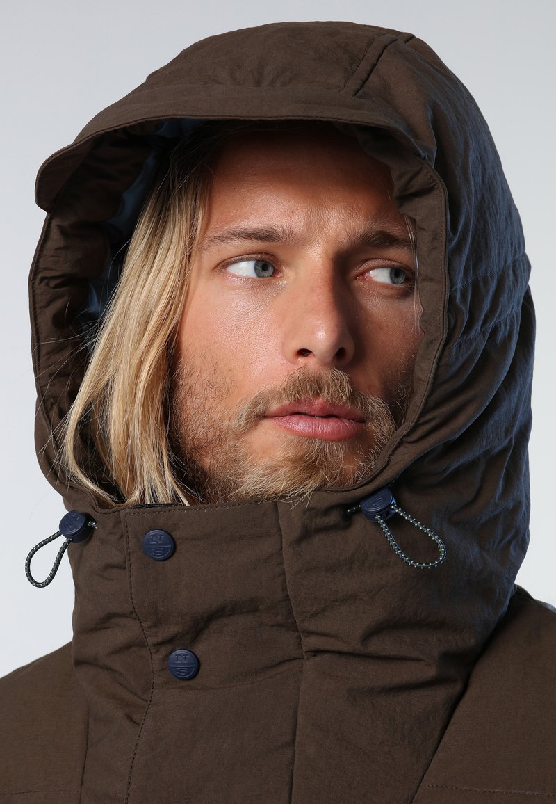 Giacca cappuccio marrone con texture trapuntata, fodera blu e regolazioni a toggle al collo. Presenta bottoni neri vicino al mento.