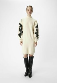 Robe en tricot couleur crème avec de longues manches brodées noires, col et poignets côtelés, associée à des bottes noires jusqu'aux genoux.