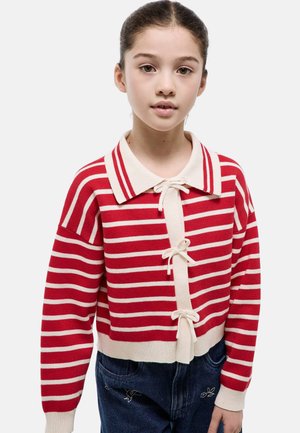 Jeune fille portant un gilet en maille rayé rouge et blanc à fermetures nœuds et un jean bleu foncé avec broderies florales.