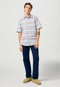Camicia a maniche corte a quadri blu con strisce rosse, dotata di una tasca sul petto. Abbinata a jeans blu scuro e scarpe mocassino color beige.