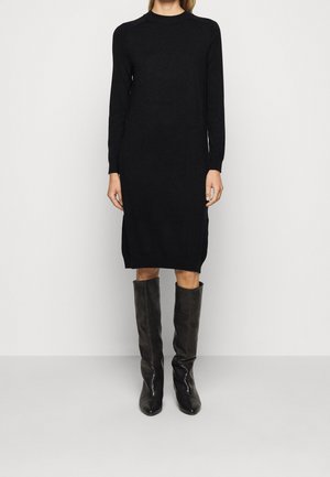 Kobieta ubrana w czarną, sweterkową sukienkę midi z długim rękawem, zestawioną z czarnymi skórzanymi kozakiem sięgającymi kolan, na jednolitym tle.