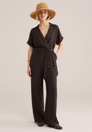 Kobieta ubrana w czarny kombinezon z paskiem, spodnie o szerokich nogawkach, słomkowy kapelusz, złota bransoletka i czarne wsuwane buty z ćwiekami, stojąca na jednolitym tle.