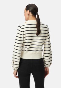 Strickpullover in Creme mit marineblauen Streifen, mit taillierter Passform und voluminösen Ärmeln. Schwarze Hosen ergänzen das Outfit.