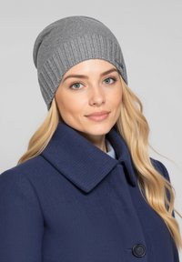 Graue Mütze mit geripptem Muster. Hellgrau, eng anliegendes Design. Getragen zu einem dunkelblauen Mantel mit rundem Kragen und einem einzelnen Knopfdetail.