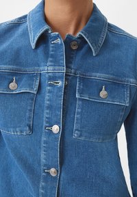 Jeansjacka i medelblått. Har en krage, två framfickor med silverknappar och en strukturerad passform. Mjuk textur med synliga sömmar.