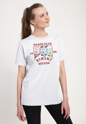 Vrouw met een wit T-shirt met kleurrijk SpongeBob-hardloopontwerp en de tekst "Track Club Bikini Bottom 1999", kijkend omhoog en opzij.