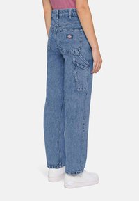 Pessoa a usar calças cargo de ganga azuis largas e sapatilhas brancas, vista de costas e cortada na cintura e nos tornozelos.