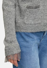 Szary sweter dziany z przednią kieszenią i ściągaczami na rękawach, z przezroczystą szarą tkaniną na wierzchu. Noszony z jasnoniebieskimi dżinsami.