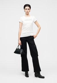 T-shirt blanc à manches courtes avec le logo "ARMANI", associée à un pantalon cargo noir et des bottines, tenant un petit sac à main noir.