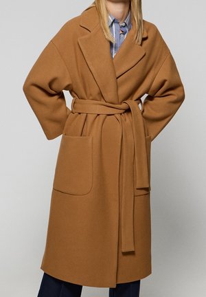 Femme portant un long manteau ceinturé camel marron avec de grands revers et des poches plaquées sur une chemise rayée bleue.