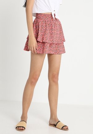 Jupe rose à couches avec un petit motif floral, taille élastique et design à volants. Portée avec des sandales à glissière noires et beiges.