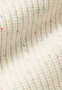 Nahaufnahme eines cremefarbenen Strickstoffs mit geripptem Muster und verstreuten kleinen farbigen Sprenkeln in Rot, Grün, Blau und Gelb.