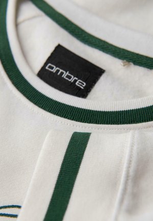 Close-up van de neklijn van een wit shirt met donkergroene trim en bijpassende groene streep op de mouw, met het zwarte merklabel "ombre."