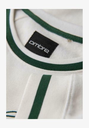 Detail bílého límce košile s tmavě zeleným lemem a odpovídající zelenou linkou na manžetě, na které je vidět černou značkovou etiketu "ombre."