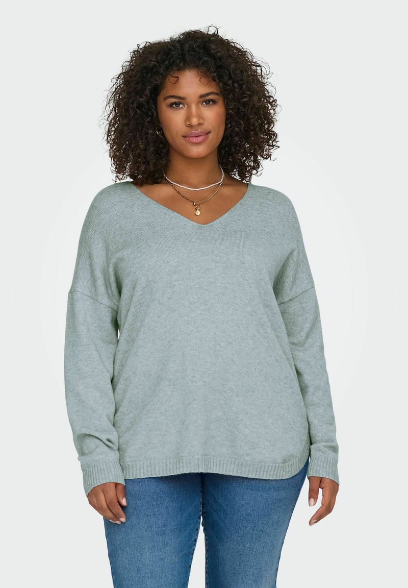 Mujer con cabello rizado, vestida con un suéter gris claro de cuello en V y jeans azules, de pie contra un fondo claro.
