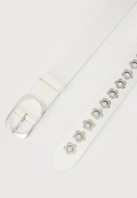 Λευκή ζώνη από συνθετικό δέρμα με ασημένιο ορθογώνιο αγκράφα, διακοσμημένη με λουλουδένια σκάλισματα στη μία πλευρά για πρόσθετη λεπτομέρεια.