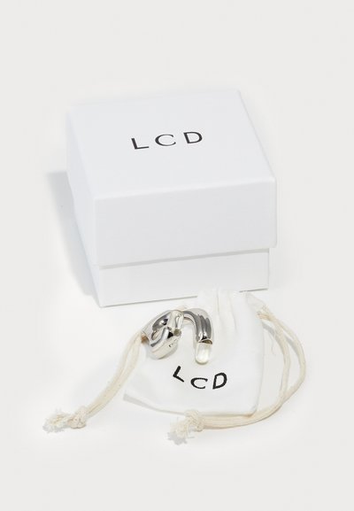 Des bijoux en métal argenté, présentant des formes courbes, rangés dans une pochette en tissu blanc portant l'inscription "LCD", à côté d'une boîte blanche avec du texte noir.