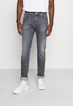 Homme portant un jean gris slim retroussé, des chaussettes blanches, des baskets blanches et un t-shirt blanc rentré, sur un fond uni.