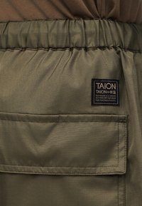 Παντελόνι με ελαστική μέση σε απόχρωση ελιάς, με μεγάλη ορθογώνια τσέπη και μαύρη ετικέτα της μάρκας TAION ραμμένη πάνω από την τσέπη.