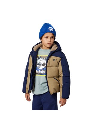 Doudoune en marine et kaki, avec une capuche, une fermeture éclair et des poches latérales. Associée à un t-shirt bleu clair et un bonnet en tricot bleu.