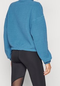 Blå fleecepolo med en texturerad yta, hög krage och elastisk nederdel. Matchas med svarta leggings som har ett genomskinligt panel.