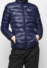 Navy puffer jacket with a high collar, quilted design, and zip closure. Features the EA7 Emporio Armani logo on the chest. Lightweight fabric.