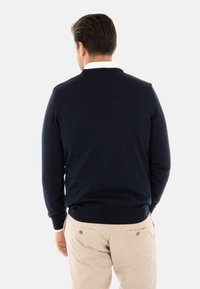 Maglione blu navy con polsini e orlo a coste, realizzato in tessuto liscio. La vista posteriore mostra un classico design a girocollo. Abbinato a pantaloni beige chiaro.