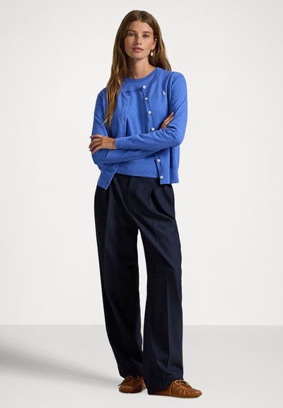 Μπλε ζακέτα με στρογγυλή λαιμόκοψη, κουμπιά στο μπροστινό μέρος και ριμπ μανσέτες, συνδυασμένη με φαρδιά μαύρα παντελόνια και ταμπά παπούτσια.