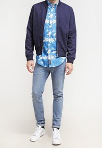Mężczyzna w granatowej kurtce bomberce, niebieskiej koszuli w wzory z guzikami, jasnoniebieskich dżinsach i białych tenisówkach stoi na jednolitym tle.