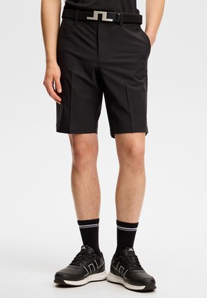 Mann trägt schwarze, maßgeschneiderte Shorts, schwarzen Gürtel, schwarze Socken mit weißen Streifen und schwarze Sneaker mit weißen Akzenten, steht vor weißem Hintergrund.